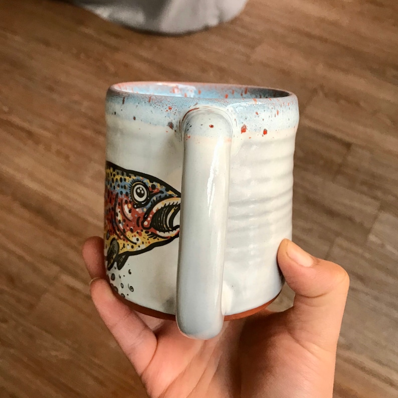 May include: White ceramic mug with a brown, red, yellow, and blue trout illustration. The mug has a speckled glaze and a brown rim.