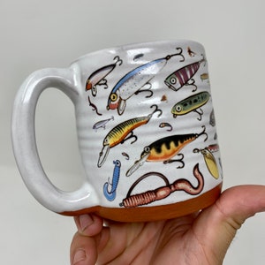 May include: White ceramic coffee mug with a colorful design of fishing lures. The mug has a rounded handle and a brown rim.