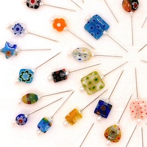 May include: A selection of decorative hat pins with vibrant glass heads in diverse shapes, such as flowers, squares, and ovals. Each pin features a silver-toned shaft and a small pearl detail. The pins are arranged in a radial pattern against a white backdrop.