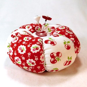 May include: A round pincushion with a red and white cherry and floral pattern. Two decorative pins with flower-shaped heads are inserted into the center. The pincushion is a sewing accessory.
