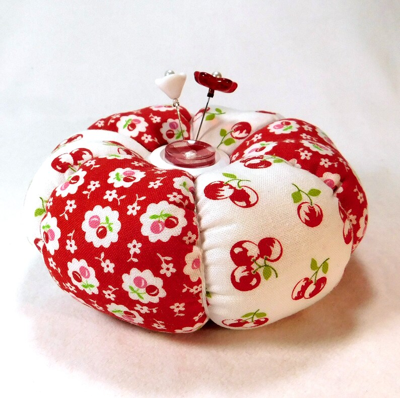 May include: A round pincushion with a red and white cherry and floral pattern. Two decorative pins with flower-shaped heads are inserted into the center. The pincushion is a sewing accessory.