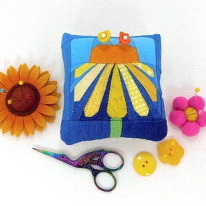 A fun, square shaped, modern pincushion featuring a bright yellow daisy on a blue ombre background. Paper pieced using a pattern by LizTaylorHandmade. 4 by 3 1/2 inches by 2 1/4 inches high. Emery core to sharpen your pins with fiberfill filling.