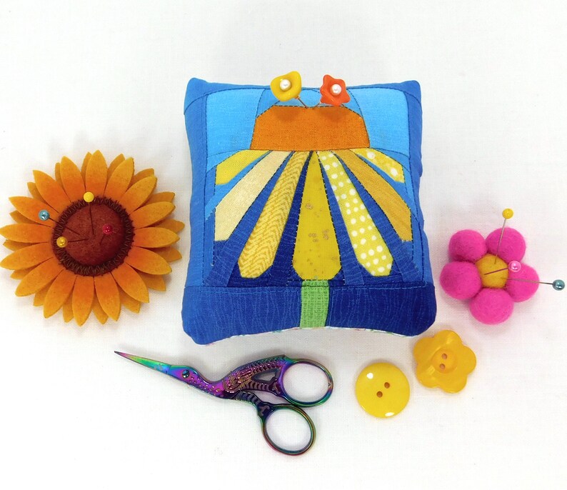 A fun, square shaped, modern pincushion featuring a bright yellow daisy on a blue ombre background. Paper pieced using a pattern by LizTaylorHandmade. 4 by 3 1/2 inches by 2 1/4 inches high. Emery core to sharpen your pins with fiberfill filling.