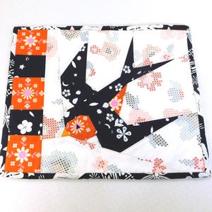 May include: A quilted, rectangular placemat featuring a black and white swallow design. The bird is set against a white background with floral and cross-stitch patterns. The border includes black, orange, and white floral blocks.