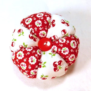 May include: A handmade, flower-shaped pin cushion. The pin cushion is made of red and white fabric with a cherry pattern. A large red button is in the center. The pin cushion is approximately 4 inches in diameter.