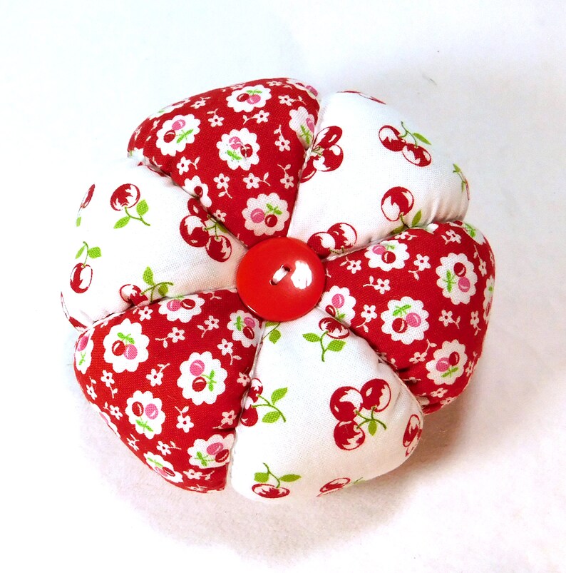 May include: A handmade, flower-shaped pin cushion. The pin cushion is made of red and white fabric with a cherry pattern. A large red button is in the center. The pin cushion is approximately 4 inches in diameter.