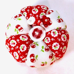May include: A handmade flower-shaped pin cushion. The pin cushion is made of red and white fabric with cherry and floral patterns. It has a large white button with a red gingham center and a small red flower with a pearl.
