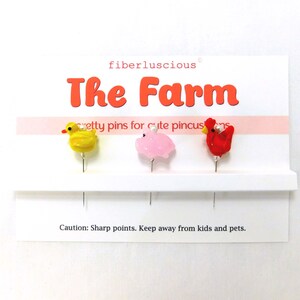 May include: A set of three farm animal pins: a yellow duck, a pink pig, and a red chicken. The pins are displayed on a white card with the text "The Farm" and "pretty pins for cute pincushions".