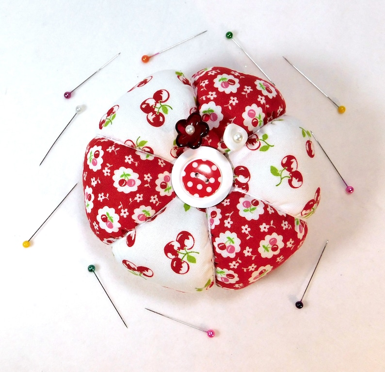 May include: A flower-shaped pin cushion with red and white cherry print fabric petals. A red and white polka dot button is in the center, surrounded by a small red flower and a white pearl. Sewing pins with colorful heads are scattered around the cushion.