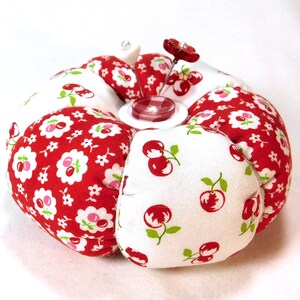 May include: A round pincushion with a white center and red fabric sections. The red fabric is patterned with white flowers and cherries. A red flower pin and a clear button are in the center. This sewing accessory is a charming addition to any craft kit.