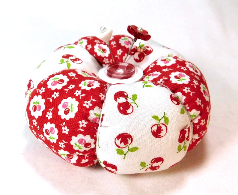 May include: A round pincushion with a white center and red fabric sections. The red fabric is patterned with white flowers and cherries. A red flower pin and a clear button are in the center. This sewing accessory is a charming addition to any craft kit.