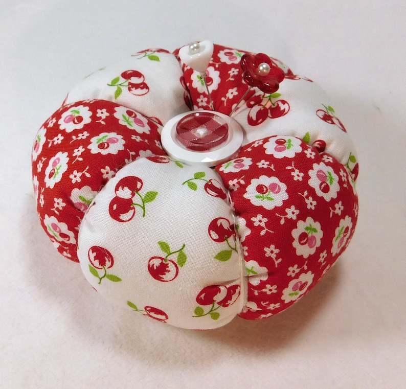 May include: A round pin cushion with a red and white cherry print fabric. The cushion is segmented into sections and features a red button and decorative pins with red flower and pearl accents. A useful sewing accessory.