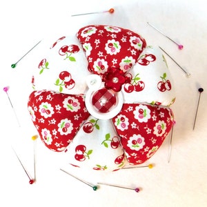 May include: A flower-shaped pin cushion with red and white cherry print fabric petals. The center features a red and white gingham button and a small red flower. Several sewing pins with colorful heads surround the cushion.