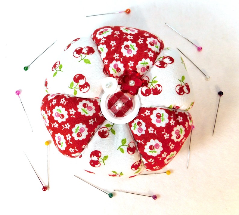 May include: A flower-shaped pin cushion with red and white cherry print fabric petals. The center features a red and white gingham button and a small red flower. Several sewing pins with colorful heads surround the cushion.