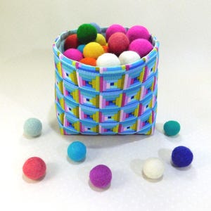 May include: A square basket with a blue, pink, and yellow geometric pattern, filled with colorful felt balls. The felt balls are various colors, including pink, green, yellow, and white. Several felt balls are scattered around the basket.