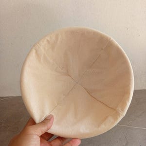 May include: A beige, circular fabric hat with a rounded crown. The hat is made of a light-colored, textured material and has a simple, minimalist design. The hat is held by a hand.