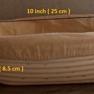 Może przedstawiać: Owalny kosz do wyrastania chleba, o długości 25 cm i wysokości 8,5 cm, z kremową podszewką z tkaniny. Kosz wykonany jest z tkanego, jasnego materiału, idealny do pieczenia chleba rzemieślniczego.