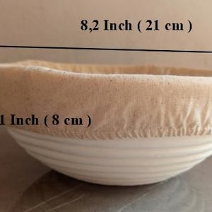 Op de afbeelding: Een ronde broodrijsmand, wit en beige. De mand is 21 cm breed en 8 cm hoog. De binnenkant is bekleed met beige stof. Ontworpen voor het rijzen van brooddeeg.