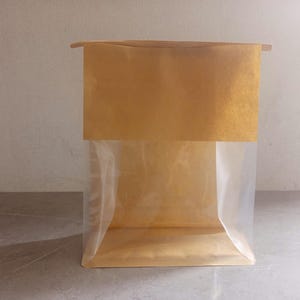 May include: A square paper bag with a clear plastic lining. The top of the bag is folded over, creating a rectangular shape. The bag is made of brown paper and clear plastic.