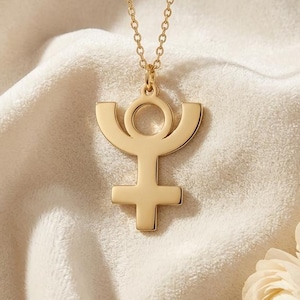 May include: A gold-colored pendant necklace featuring a stylized symbol with a circular top, curved arms, and a cross at the bottom. The necklace hangs on a delicate chain against a soft, cream-colored fabric. Two cream-colored flowers are in the lower right corner.