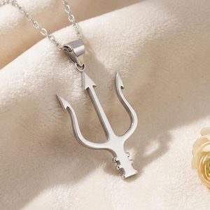 A polished silver Neptune Trident Necklace featuring a three-pronged spear design, symbolizing power and the ocean. The mythological pendant hangs from a delicate silver cable chain, resting on a light beige surface next to white decorative stones and dried natural elements. This ocean symbol jewelry piece is captured in a bright, soft-lit setting, showcasing its smooth, reflective surface and intricate details.