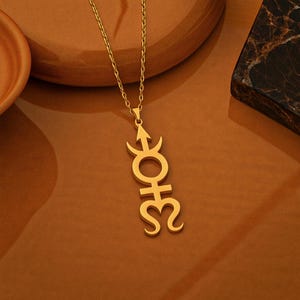 May include: A gold-colored pendant necklace featuring a stylized symbol. The pendant hangs from a delicate chain and is displayed against a warm brown background. The symbol has an arrow, a circle, a cross, and a curved line.