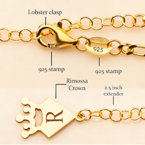 May include: Close-up of a gold-colored necklace with a lobster clasp and a 2.5-inch extender. The necklace features a crown pendant with the letter "R" and a "925" stamp. The chain is made of small, connected links.