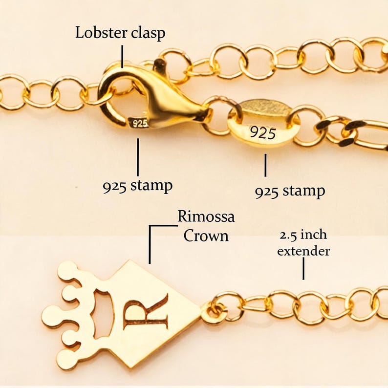 May include: Close-up of a gold-colored necklace with a lobster clasp and a 2.5-inch extender. The necklace features a crown pendant with the letter "R" and a "925" stamp. The chain is made of small, connected links.
