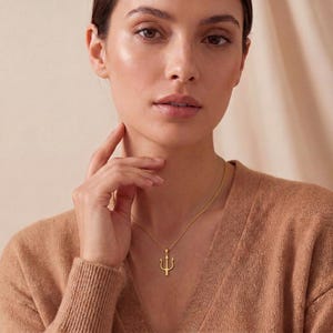 May include: A gold-colored necklace with a trident pendant. The pendant is a three-pronged fork design, suspended from a delicate chain. The necklace is worn by a person wearing a tan sweater.