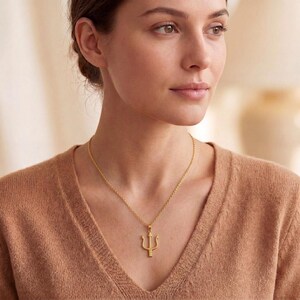 May include: A gold-colored necklace featuring a trident pendant. The pendant is a three-pronged fork design, suspended from a delicate chain. The necklace is worn against a brown sweater.