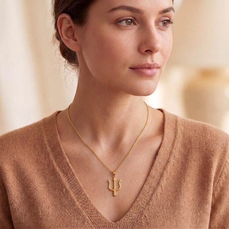 May include: A gold-colored necklace featuring a trident pendant. The pendant is a three-pronged fork design, suspended from a delicate chain. The necklace is worn against a brown sweater.