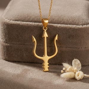 May include: A gold-colored trident pendant necklace. The pendant features a three-pronged design with an arrow-shaped center. The necklace is displayed on a gray velvet surface, with a small floral arrangement in the background.