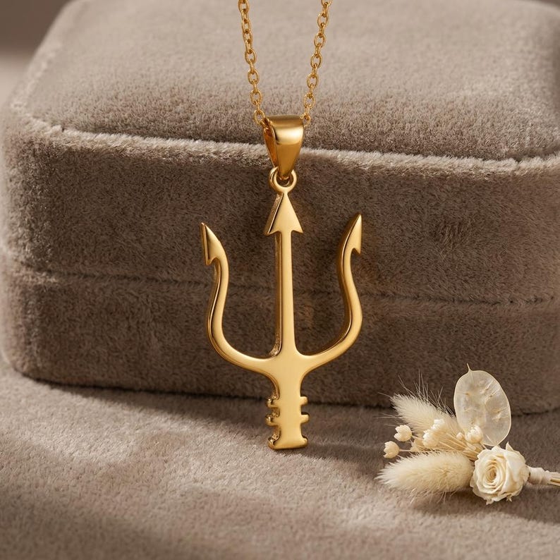 May include: A gold-colored trident pendant necklace. The pendant features a three-pronged design with an arrow-shaped center. The necklace is displayed on a gray velvet surface, with a small floral arrangement in the background.
