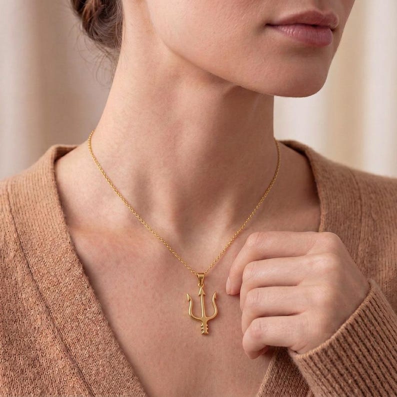 May include: A gold-colored trident pendant necklace. The necklace features a delicate chain and a detailed trident charm. The trident is a three-pronged spear, a symbol of power and authority. The necklace is worn around the neck.