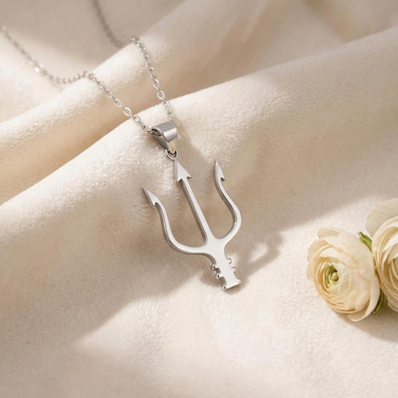A polished silver Neptune Trident Necklace featuring a three-pronged spear design, symbolizing power and the ocean. The mythological pendant hangs from a delicate silver cable chain, resting on a light beige surface next to white decorative stones and dried natural elements. This ocean symbol jewelry piece is captured in a bright, soft-lit setting, showcasing its smooth, reflective surface and intricate details.