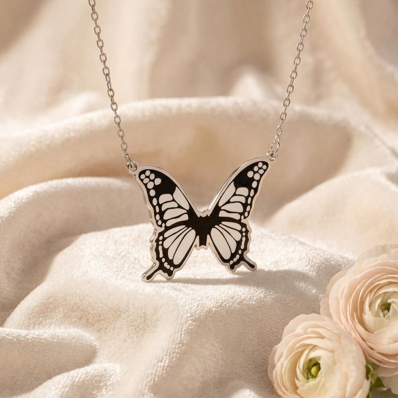 A highly detailed 925 sterling silver Monarch Butterfly Necklace with a polished finish, featuring intricate wing patterns in black enamel, rests on a light-colored fabric surface. The elegant nature-inspired pendant, symbolizing transformation and freedom, is centrally positioned on a delicate cable chain, reflecting a thoughtful gift for women. The butterfly's wings are spread open, showcasing the distinctive white dots and lines against the dark background, with a subtle reflection on the