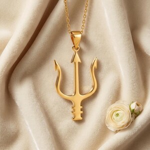 May include: A gold-colored trident pendant necklace. The pendant has a three-pronged design with an arrow-shaped center. The necklace is displayed on a cream-colored fabric, with a white flower in the lower right corner.