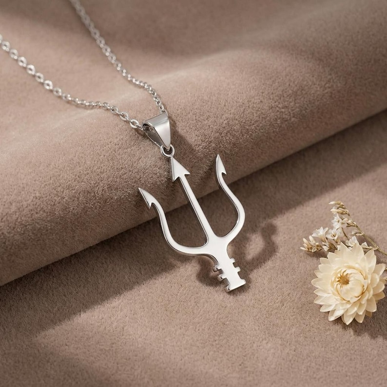 May include: A silver-colored pendant necklace featuring a trident design. The pendant is suspended from a delicate chain. The trident has three prongs and an arrow-shaped central element. The necklace is displayed on a neutral-toned surface.