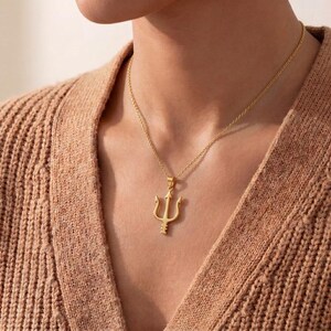 May include: A gold-colored necklace featuring a trident pendant. The pendant is a three-pronged fork design, suspended from a delicate chain. The necklace is worn against a person's skin, and the background is a tan-colored sweater.