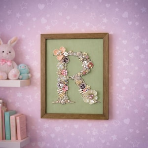 Puede incluir: Una obra de arte enmarcada que muestra la letra "R", compuesta por varios adornos florales. La letra está sobre un fondo verde claro dentro de un marco de madera. La pared detrás del marco es de color rosa claro.