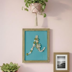 Puede incluir: Una composición decorativa de pared con una obra de arte turquesa enmarcada con un diseño floral de la letra "A", una pequeña foto en blanco y negro enmarcada y una planta colgante en una maceta rosa. Debajo hay una suculenta en maceta.