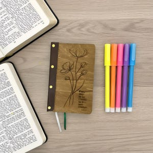May include: A wooden journal with a floral design and the quote "If any mean be in Christ, he is a new creature." Next to it are six colorful highlighters and two open books.