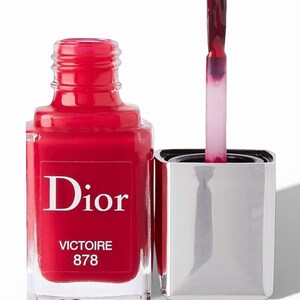 May include: A bottle of Dior nail polish in a vibrant red shade, with the name "Victoire 878" printed on the label. The bottle is open, with the applicator brush dipped in the polish, ready for use. The bottle has a silver cap.