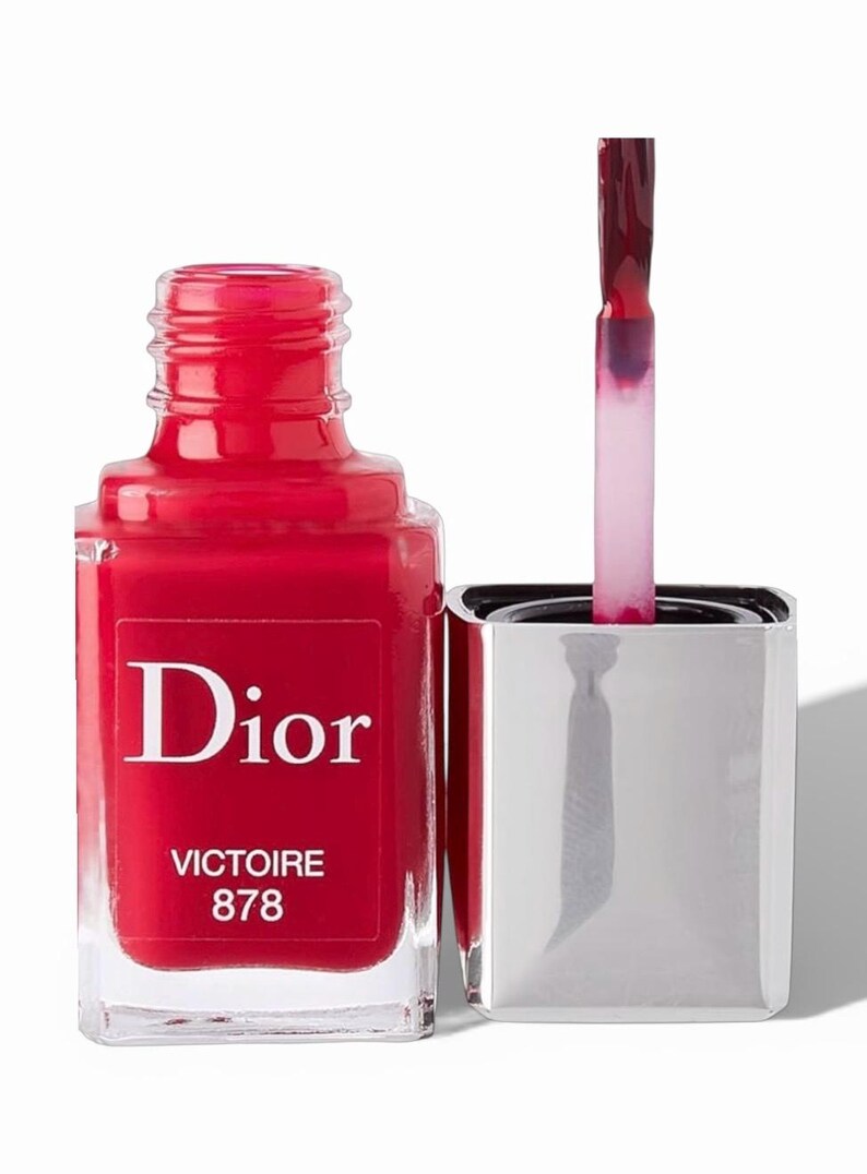 May include: A bottle of Dior nail polish in a vibrant red shade, with the name "Victoire 878" printed on the label. The bottle is open, with the applicator brush dipped in the polish, ready for use. The bottle has a silver cap.