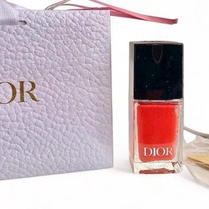 May include: A bottle of red nail polish with a black cap, a white gift bag, and a gold star-shaped charm. The gift bag and nail polish bottle both have the word "DIOR" printed on them. The gift bag has a white ribbon.