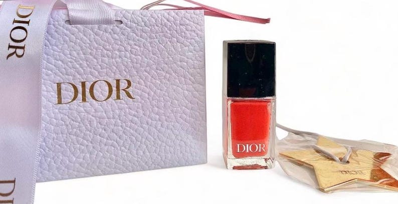 May include: A bottle of red nail polish with a black cap, a white gift bag, and a gold star-shaped charm. The gift bag and nail polish bottle both have the word "DIOR" printed on them. The gift bag has a white ribbon.