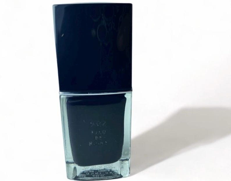 May include: A dark navy blue nail polish bottle with a rectangular shape and a matching cap. The bottle is on a white background, casting a shadow to the right. The nail polish is a deep, opaque color.