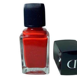 May include: A bottle of red nail polish with a black cap and a separate black cap with a silver CD logo. The nail polish bottle is square with a clear glass body and a dark red polish color.