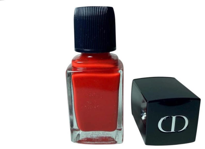 May include: A bottle of red nail polish with a black cap and a separate black cap with a silver CD logo. The nail polish bottle is square with a clear glass body and a dark red polish color.