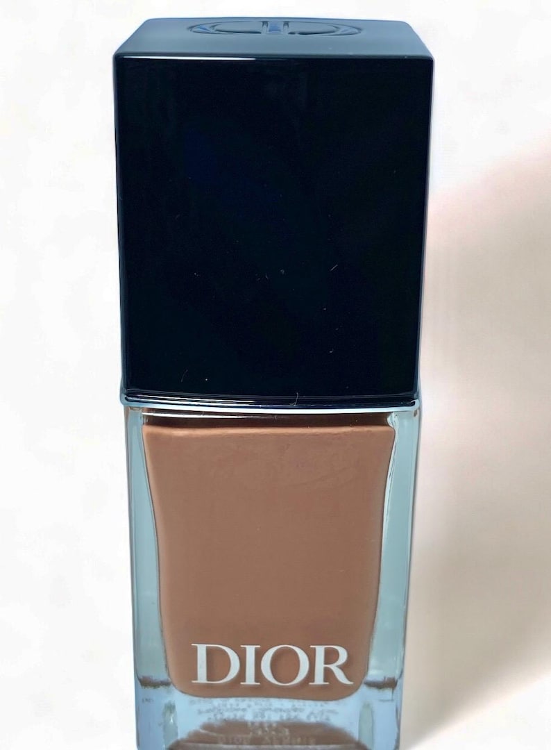 Peut inclure: Un flacon de vernis &agrave; ongles Dior. Le flacon est rectangulaire avec un bouchon noir et un corps en verre transparent. La couleur du vernis &agrave; ongles est un marron chaud. Le mot "DIOR" est imprim&eacute; en blanc sur le devant du flacon.